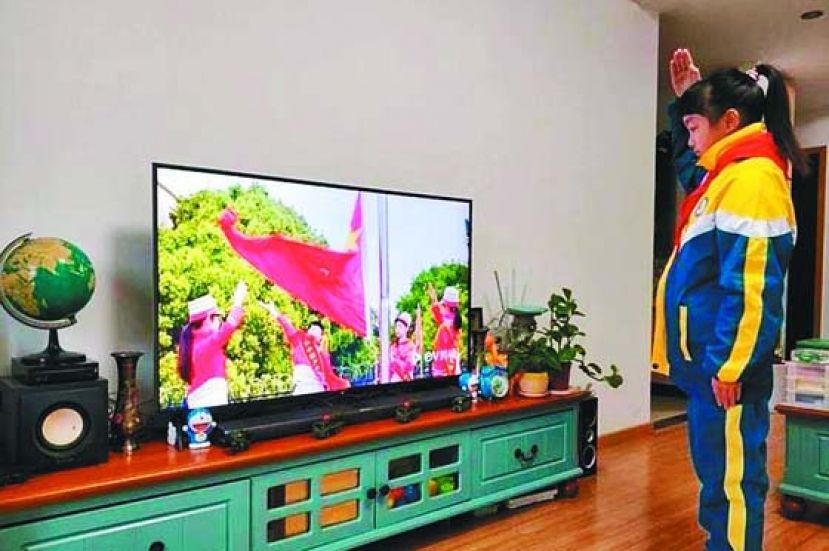 学生笔挺地站在家里电视屏幕前，胸口系著红领巾，朝著电视屏幕中的五星红旗敬礼。
